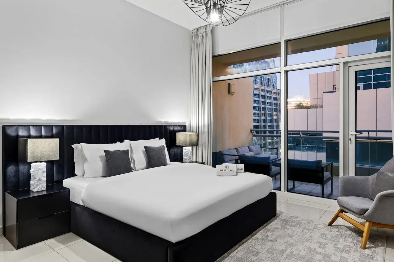 A bedroom with a neatly made bed and large windows overlooking nearby buildings.