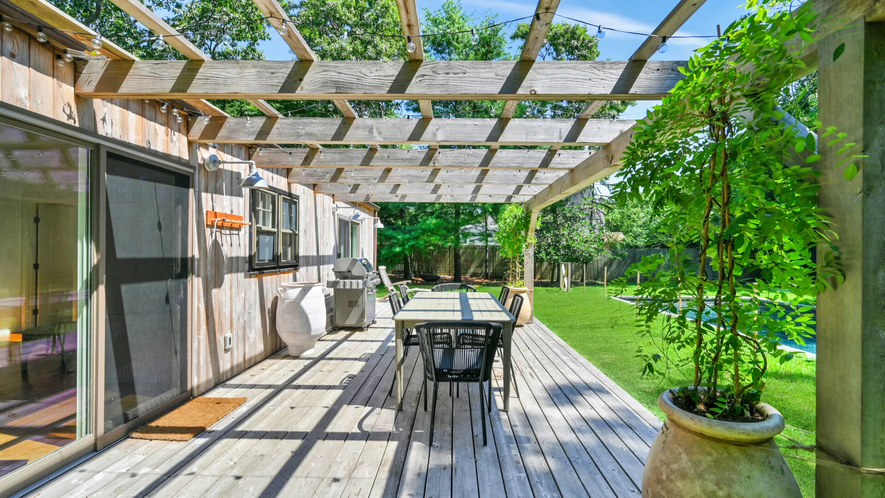 An outdoor dining area beneath a wooden pergola, with a table and surrounding greenery.