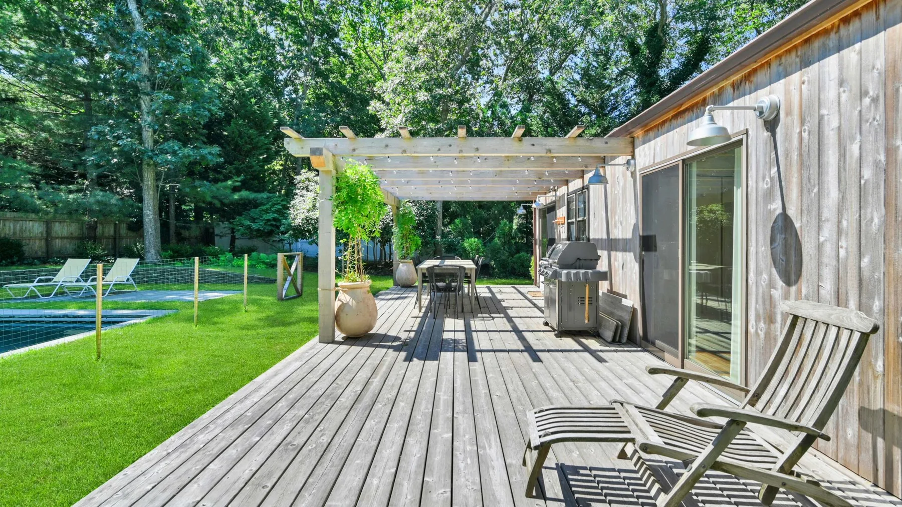 A wooden deck beside a house has a pergola, outdoor seating, and a lawn.