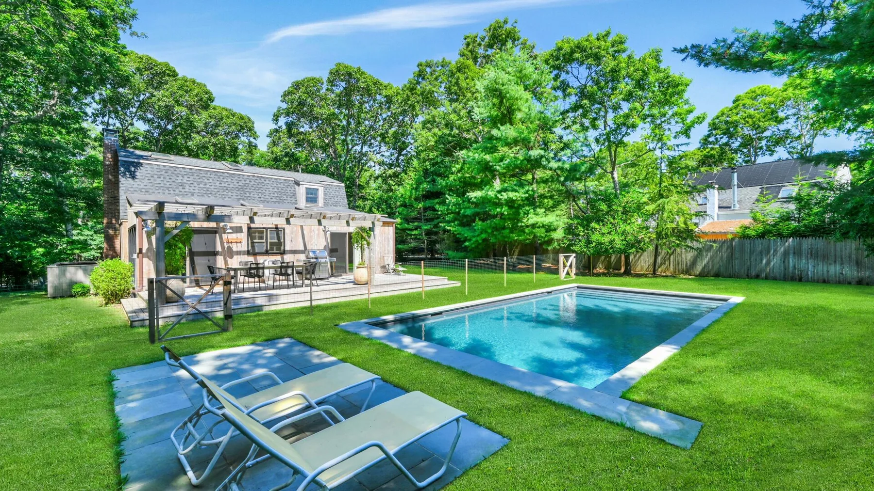 An outdoor swimming pool sits beside a grassy yard and a patio with seating.