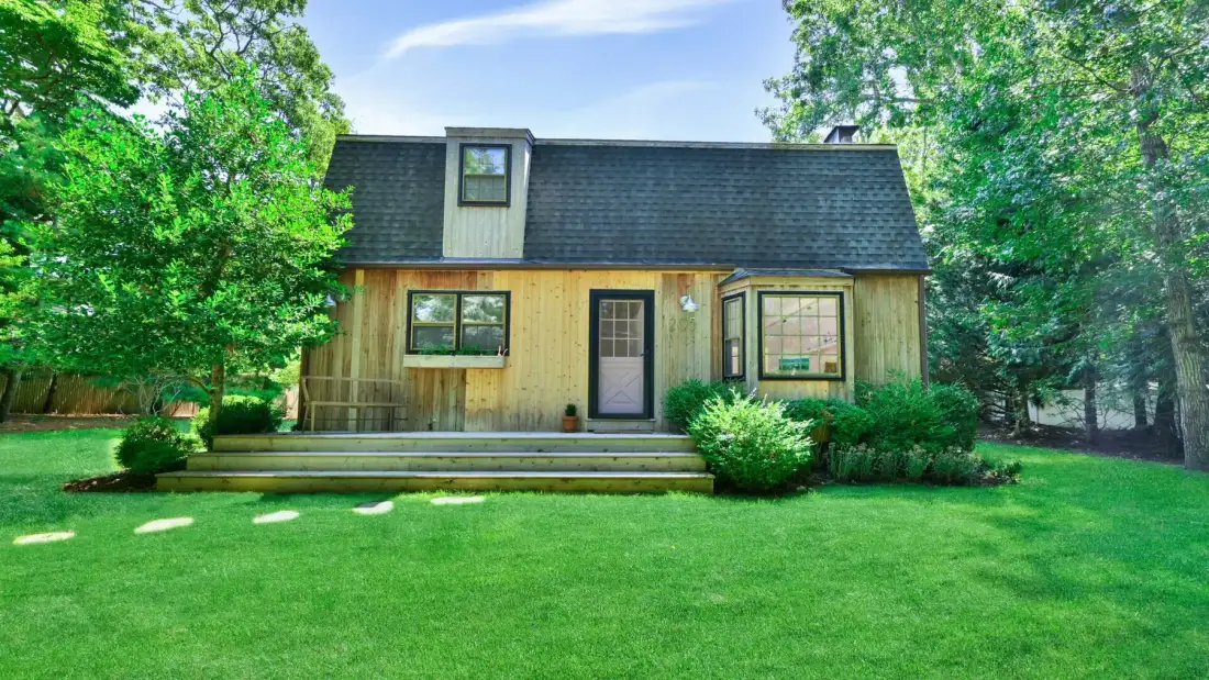 A wooden house with a dark roof stands beside a green lawn and trees.
