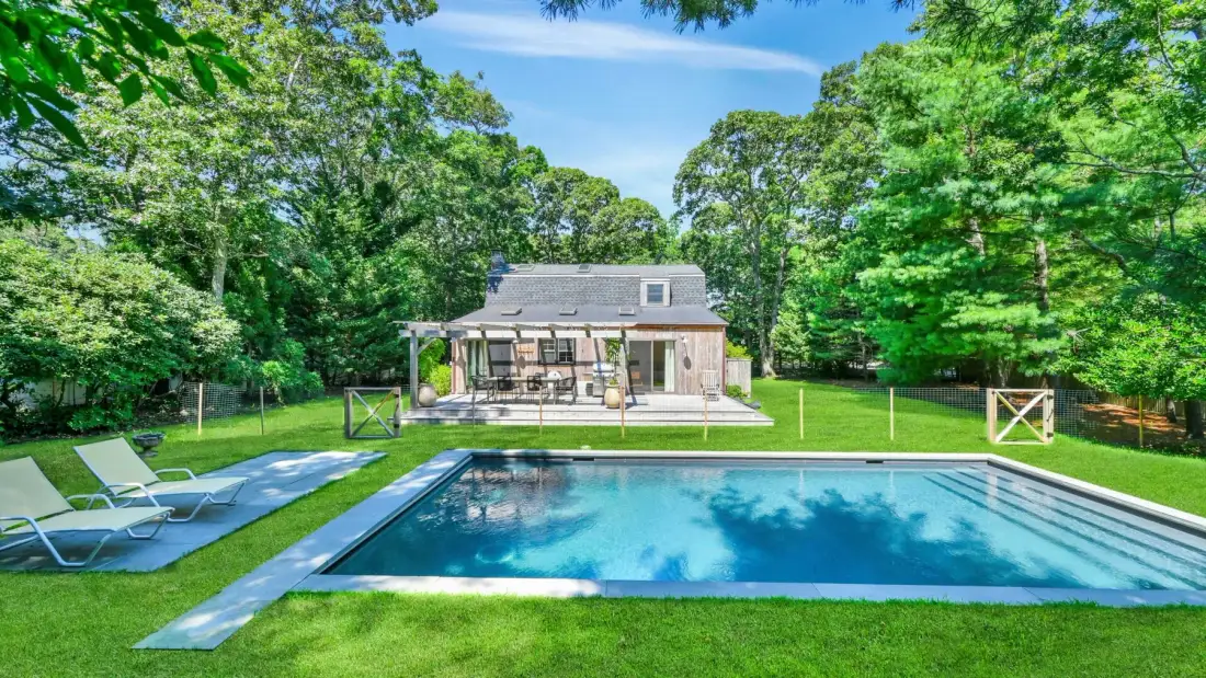 A backyard swimming pool sits in front of a house surrounded by trees.