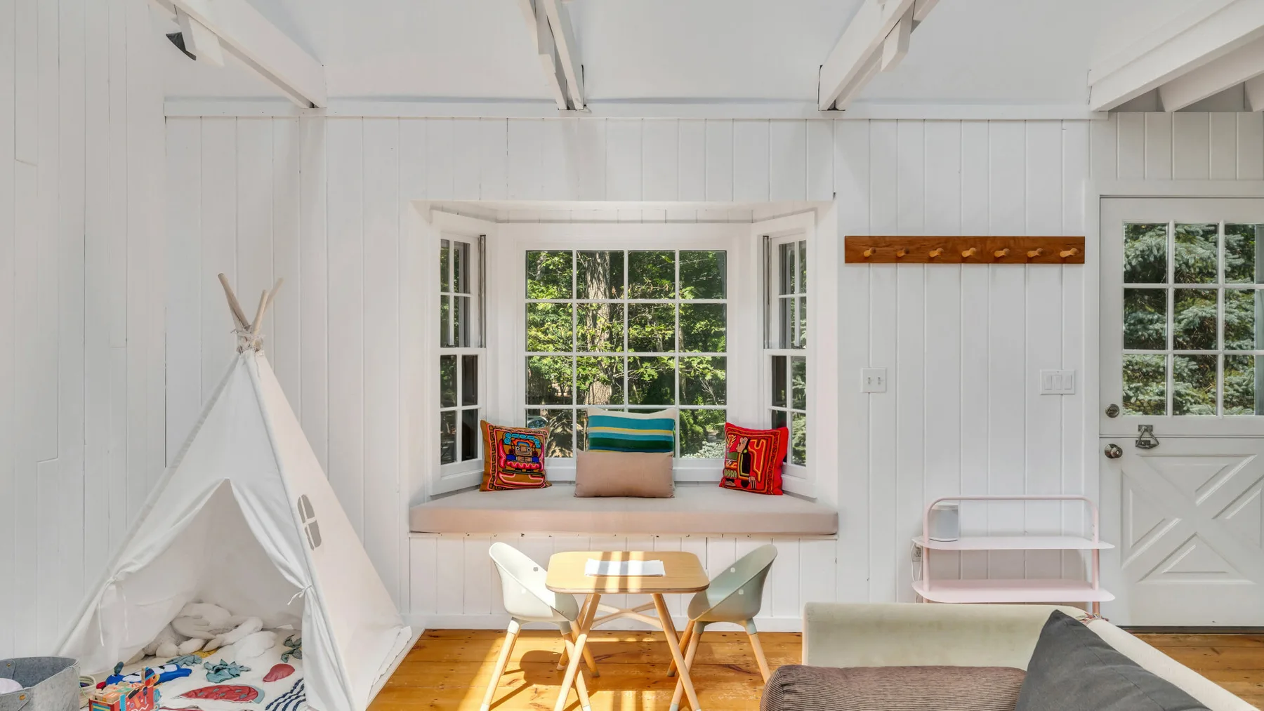 A bright white playroom has a window seat, small table, and fabric tent.