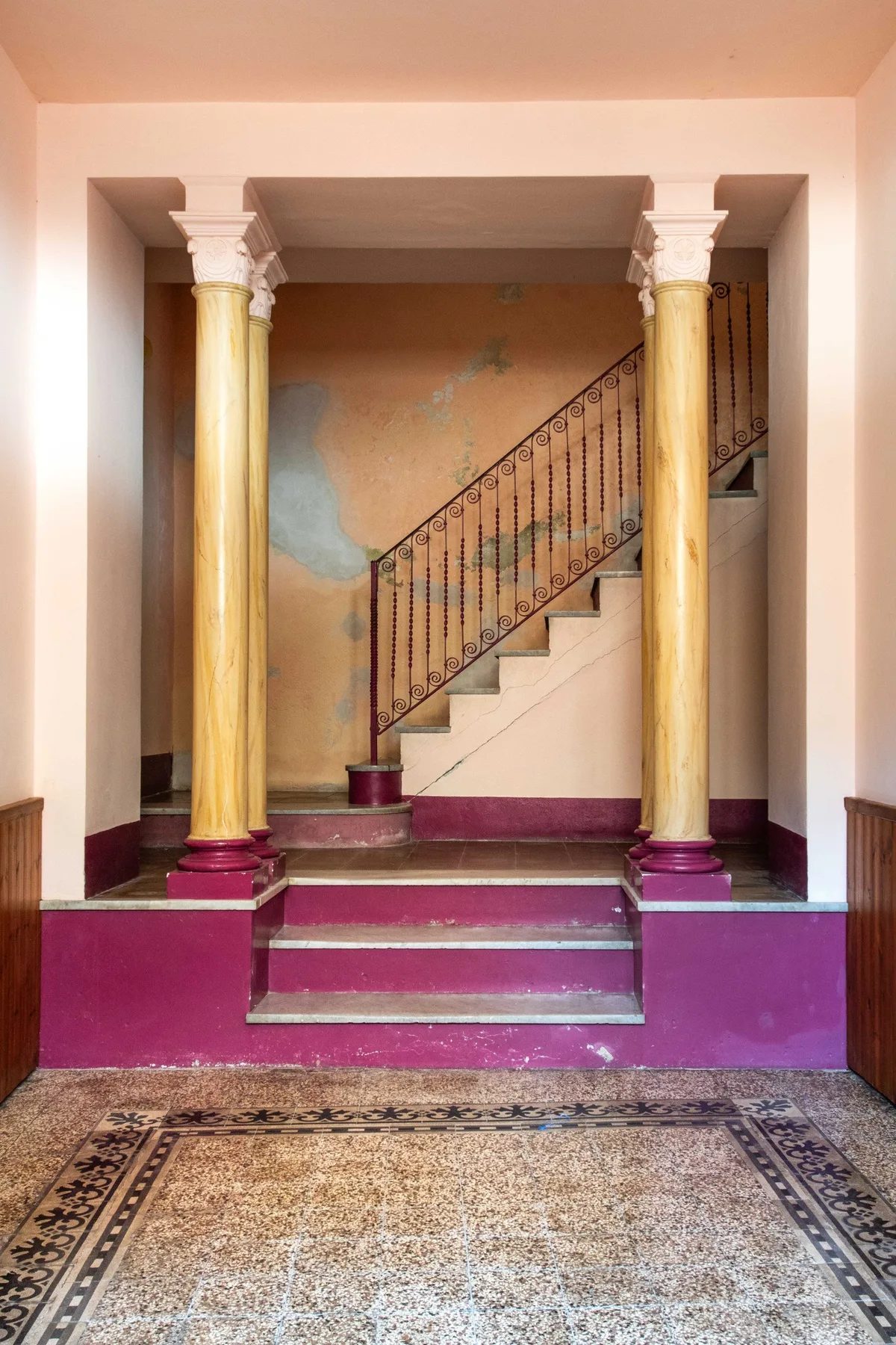 An interior stairwell rises behind decorative columns, with a patterned rug on the floor.