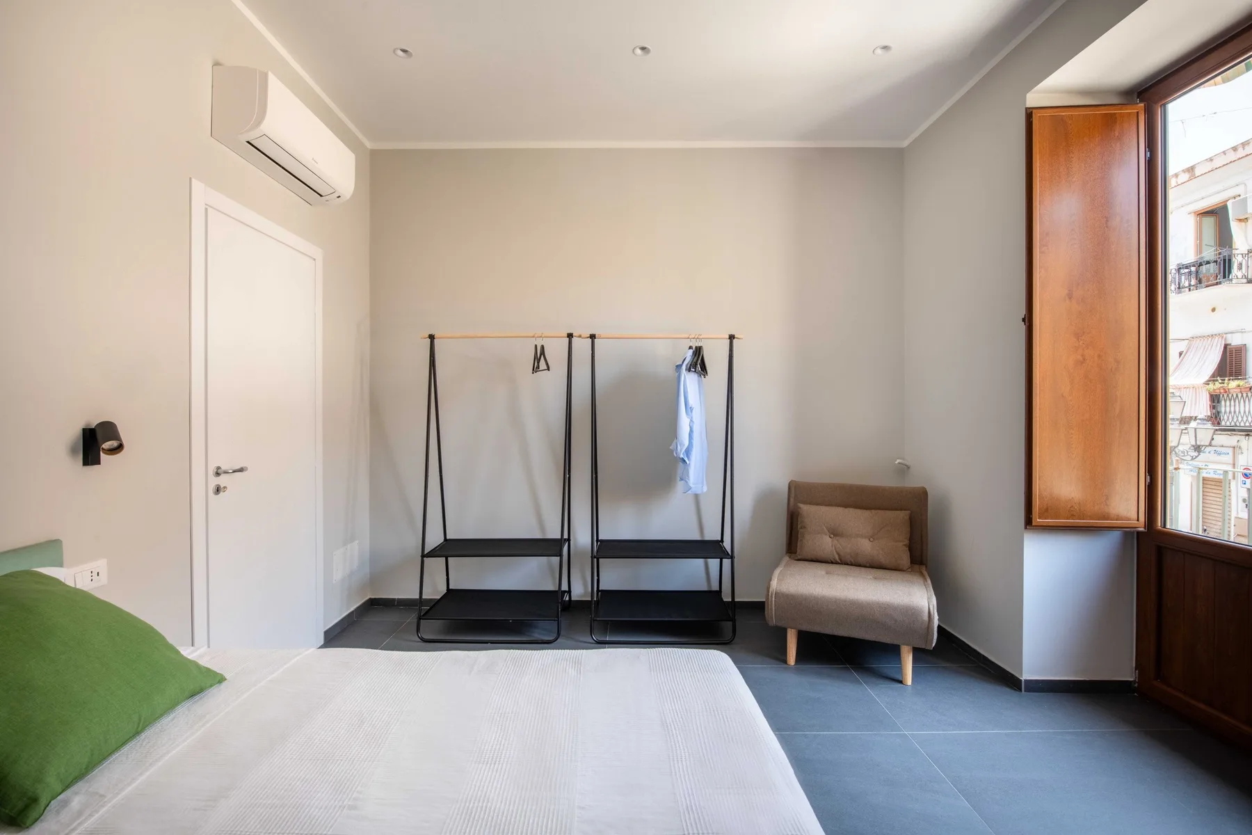 A bright bedroom with a bed, clothing racks, and a small upholstered seat.