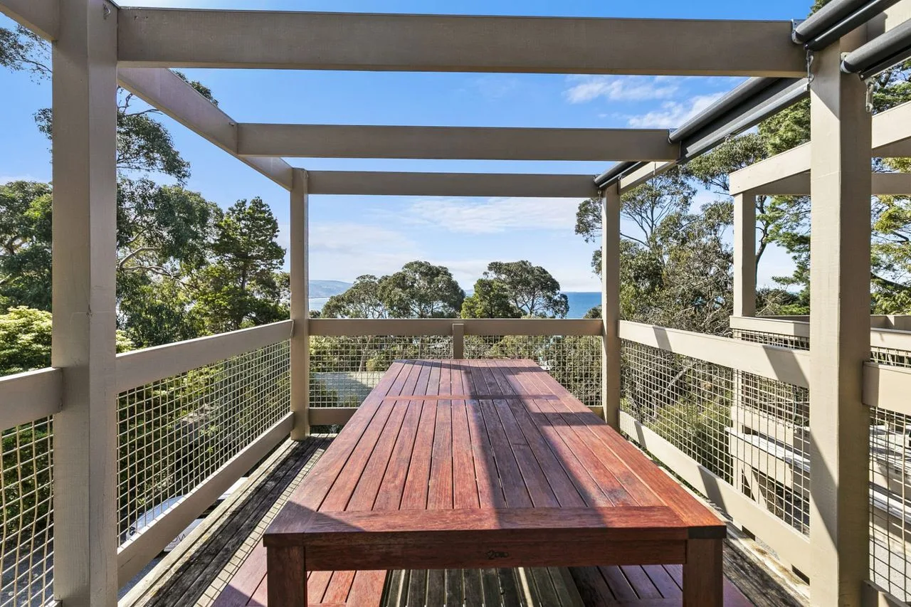 An outdoor deck with a long wooden table overlooks trees and a glimpse of the ocean.