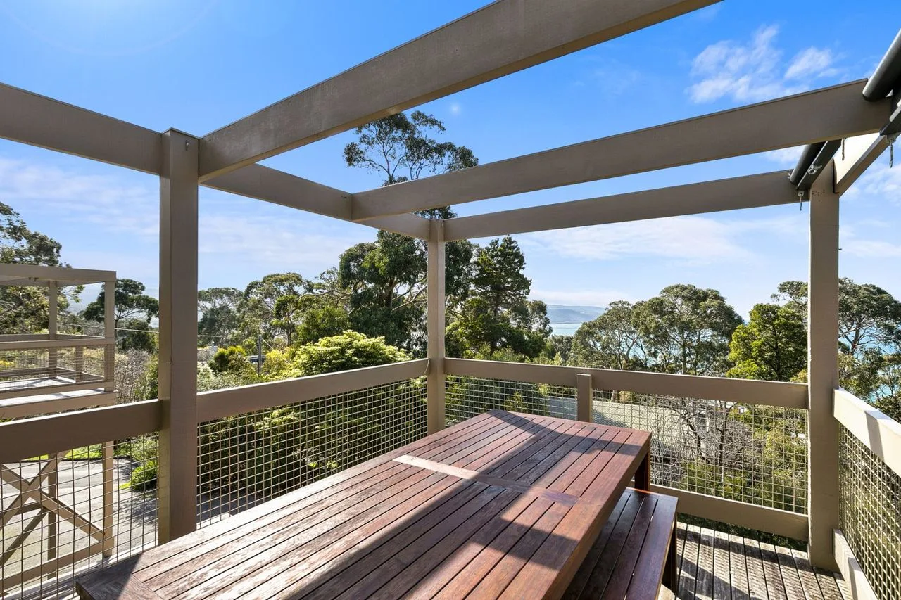 A wooden outdoor deck with a picnic table overlooks trees and a glimpse of water.
