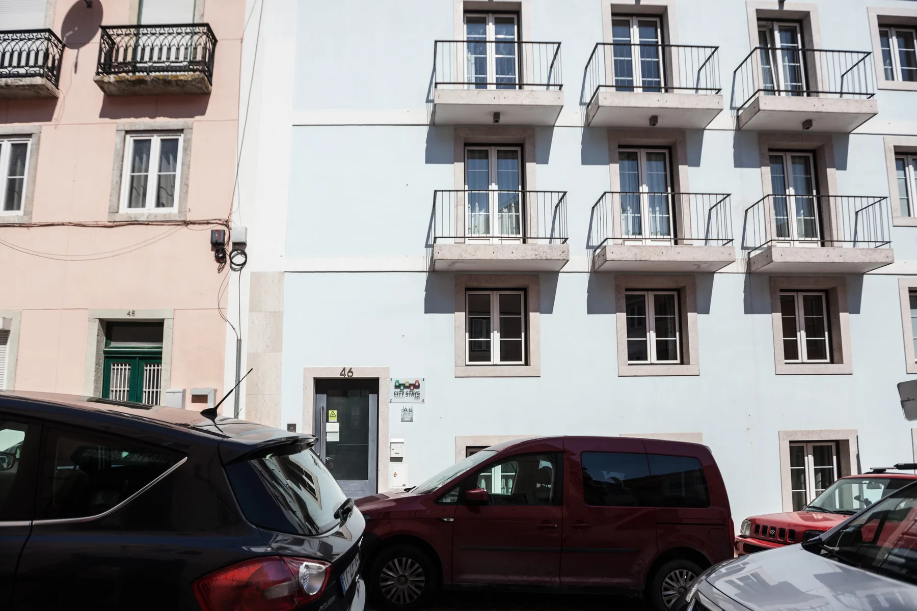 Una fachada de edificio claro con balcones pequeños da a una calle con coches aparcados.