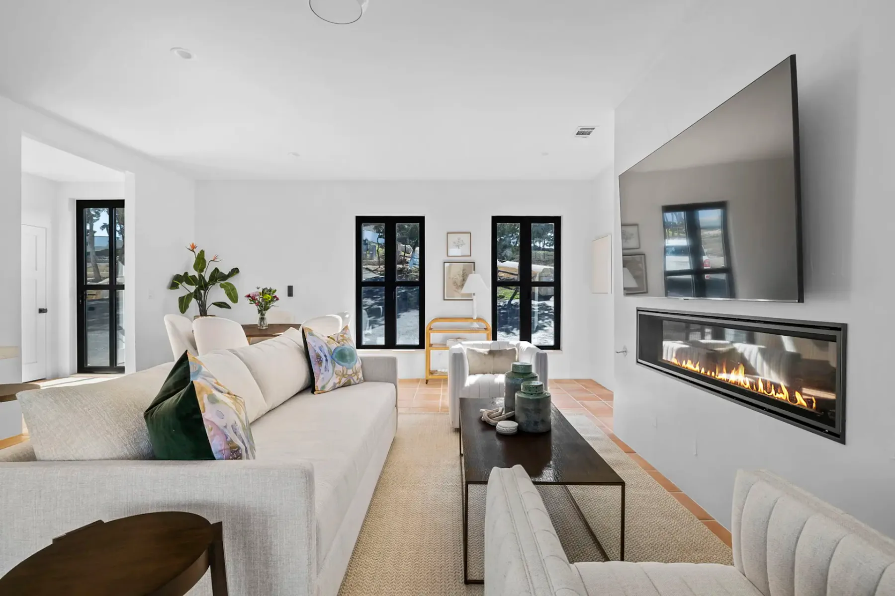 Modern living room with a large wall-mounted TV, sleek linear fireplace, beige sofas, wooden coffee table, and large windows with black frames.