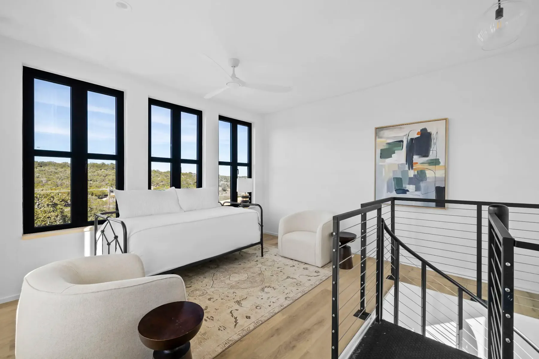 Minimalist living space with white sofa, modern chairs, abstract artwork, large windows, and a staircase with cable railing.