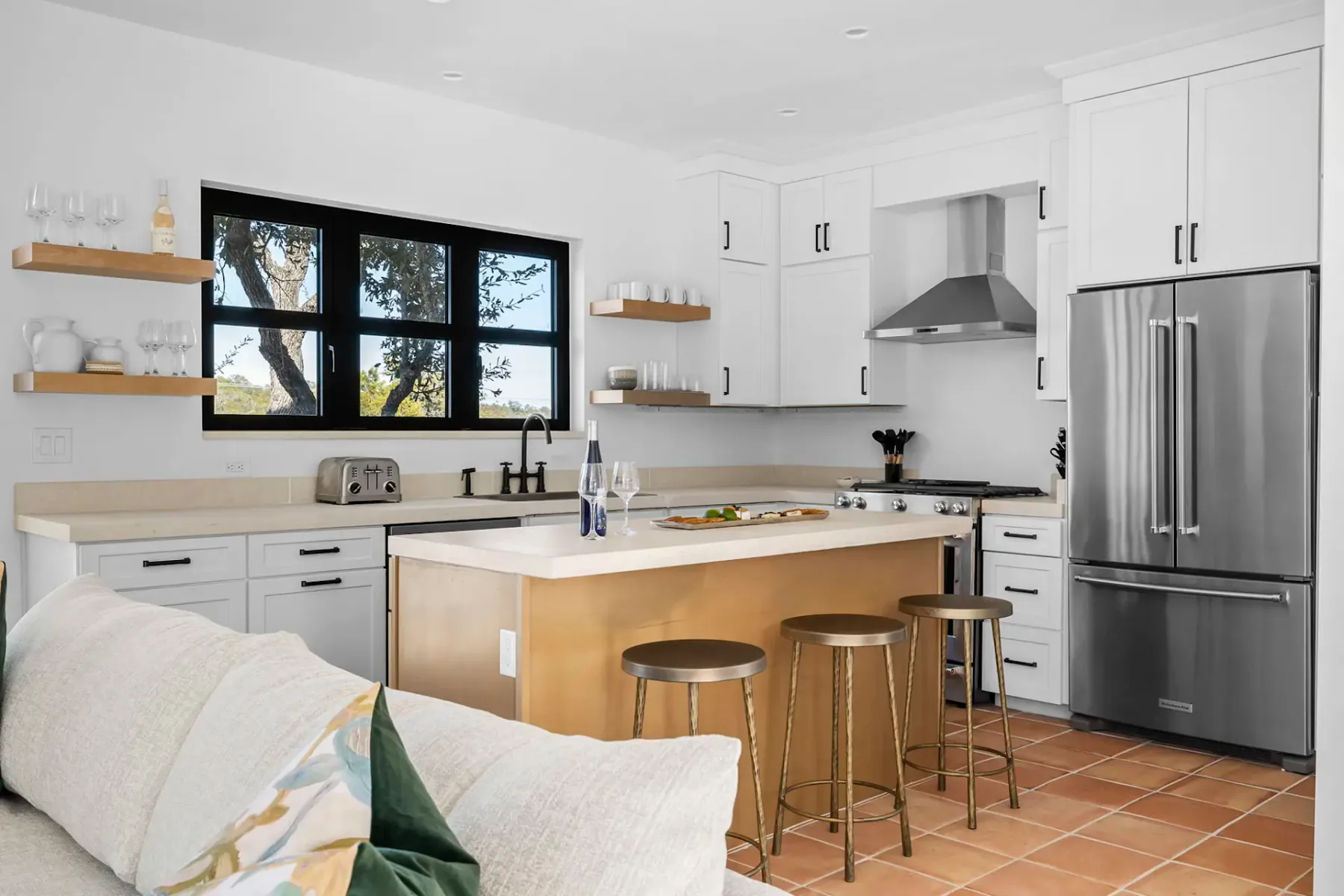 Modern kitchen with white cabinets, stainless steel appliances, a central island with stools, open shelves, and a window with a tree view.