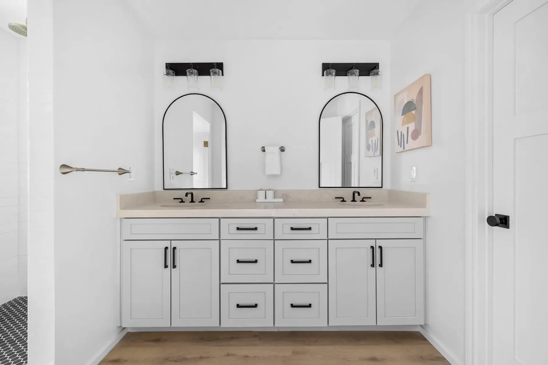 Modern bathroom with a double-sink vanity, arched mirrors, light fixtures, and neutral decor. Wooden floor and abstract wall art are visible.