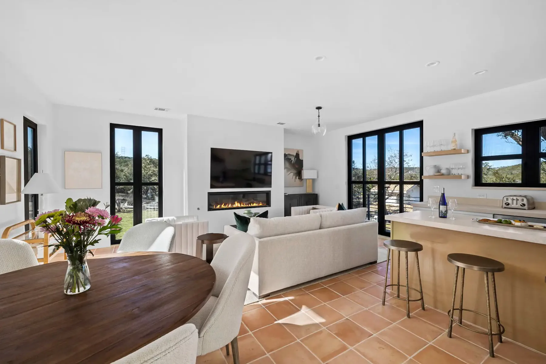 Modern living room with a cozy seating area, wall-mounted TV, fireplace, and a dining table. Large windows offer natural light and scenic views.