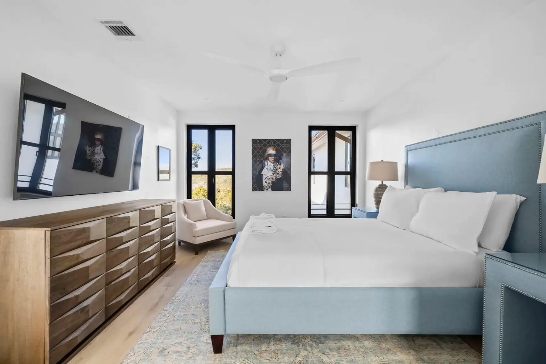A bright bedroom with a large bed, a dresser, wall art, and black-framed windows.