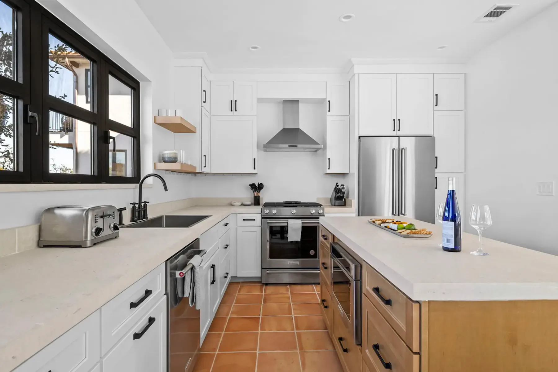 Modern kitchen with white cabinets, stainless steel appliances, black window, and terracotta floor tiles. Wine bottle and glasses on the island.