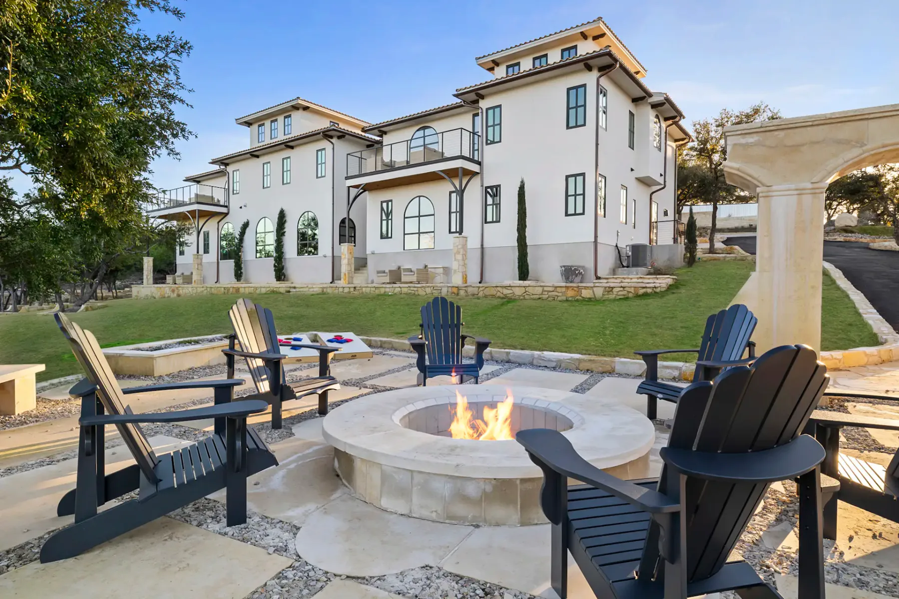 Luxurious multi-story house with large windows, surrounded by a grass lawn, features a stone patio with a fire pit and Adirondack chairs.