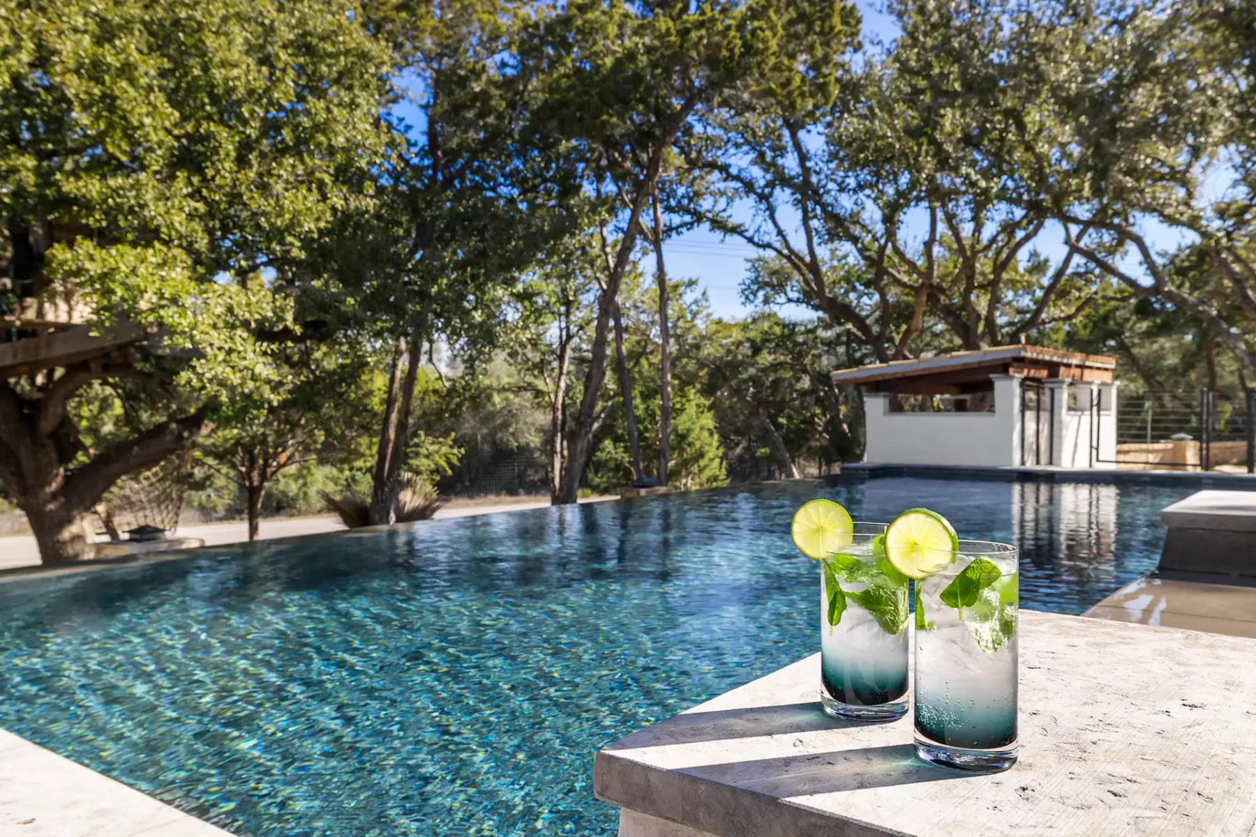 A tree-lined swimming pool with two mint and lime drinks on the edge.