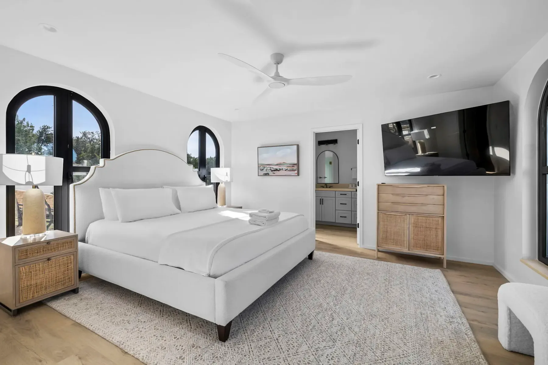 Bright bedroom with a large bed, white linens, wooden nightstands, ceiling fan, wall-mounted TV, and a doorway leading to a bathroom.