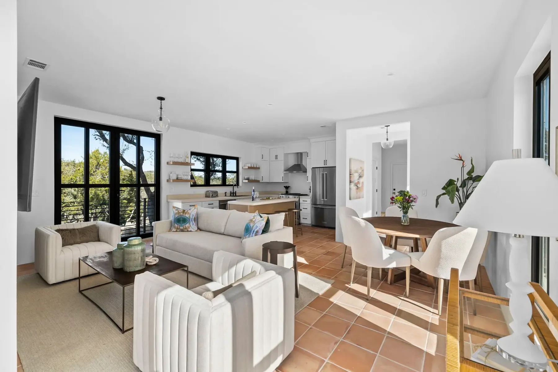 Modern open-plan living area with beige furniture, a round dining table, and large windows overlooking a green landscape. White walls and terracotta tiles.