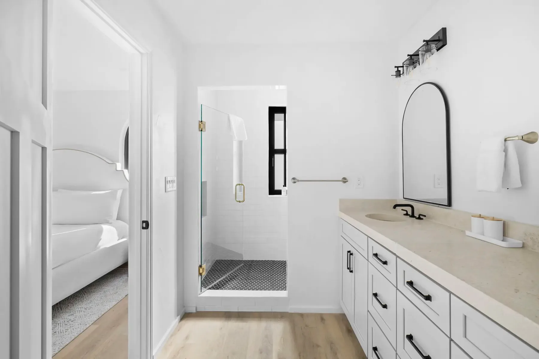 A bright bathroom with a long vanity, arched mirror, and shower; a bedroom is visible through the doorway.