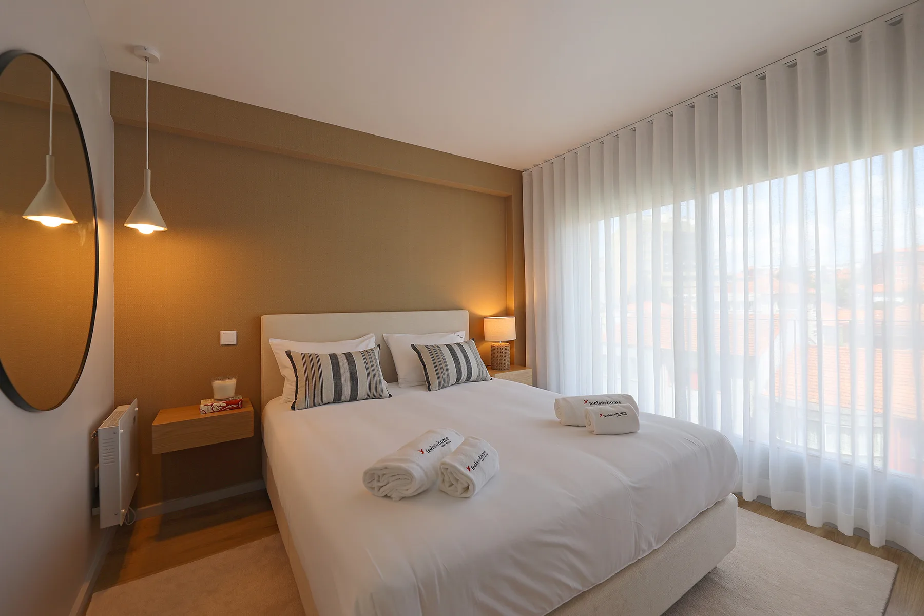 A bedroom with a neatly made bed beside a large window with sheer curtains.