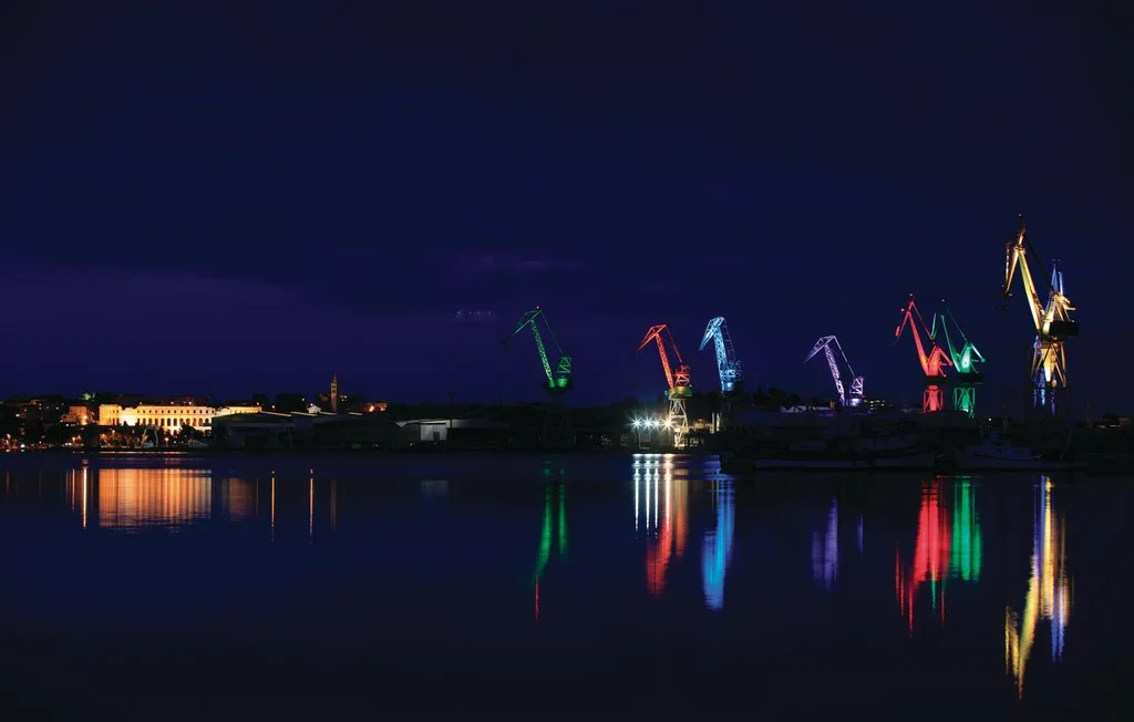 Una escena nocturna junto al agua muestra grúas iluminadas de colores reflejadas en el agua.
