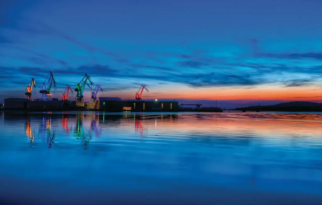 Un puerto al anochecer, con grúas reflejadas en el agua tranquila.