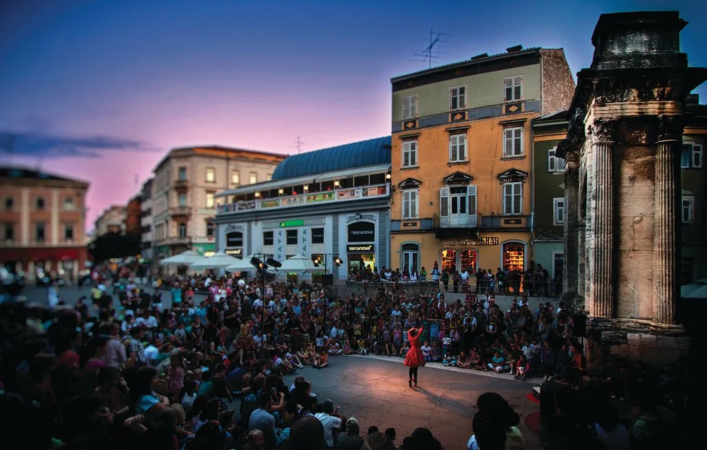 Una plaza al aire libre llena de gente, con una artista frente a edificios antiguos.