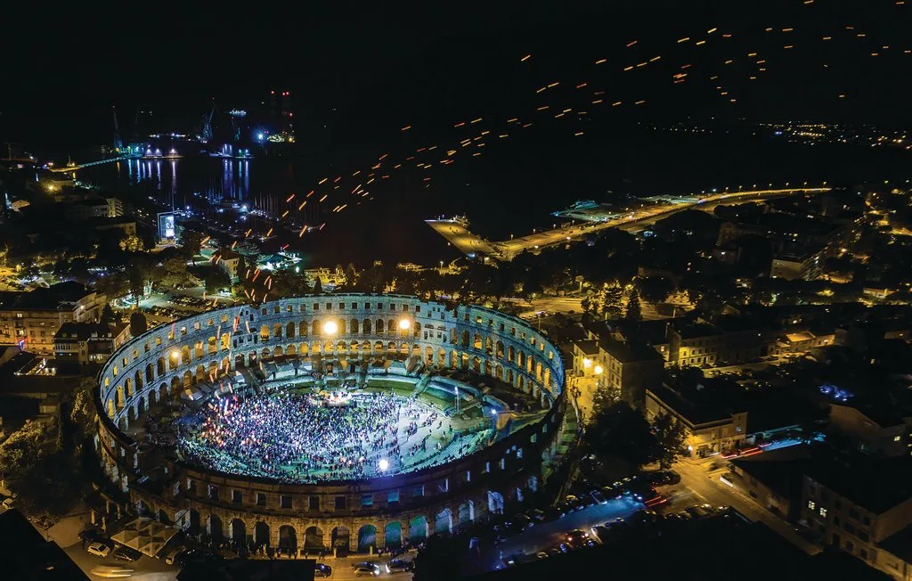Vista aérea nocturna de un anfiteatro iluminado, rodeado de luces de la ciudad.