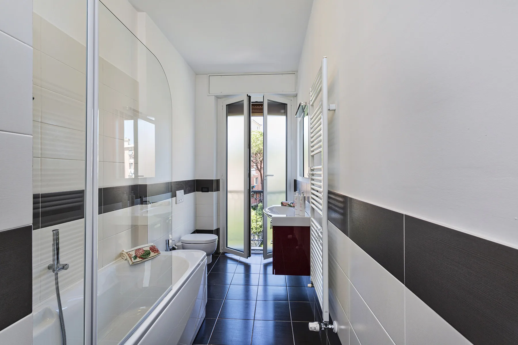 Un baño luminoso con bañera y una puerta de cristal estrecha abierta a un balcón.
