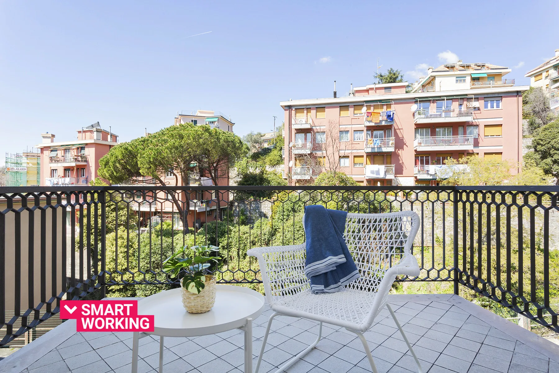 Un balcón embaldosado tiene asientos blancos, una mesa pequeña y vistas a edificios cercanos.