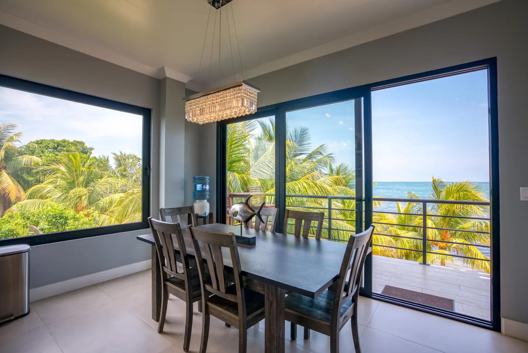 A dining area with a table and chairs beside large windows overlooking palm trees and the ocean.