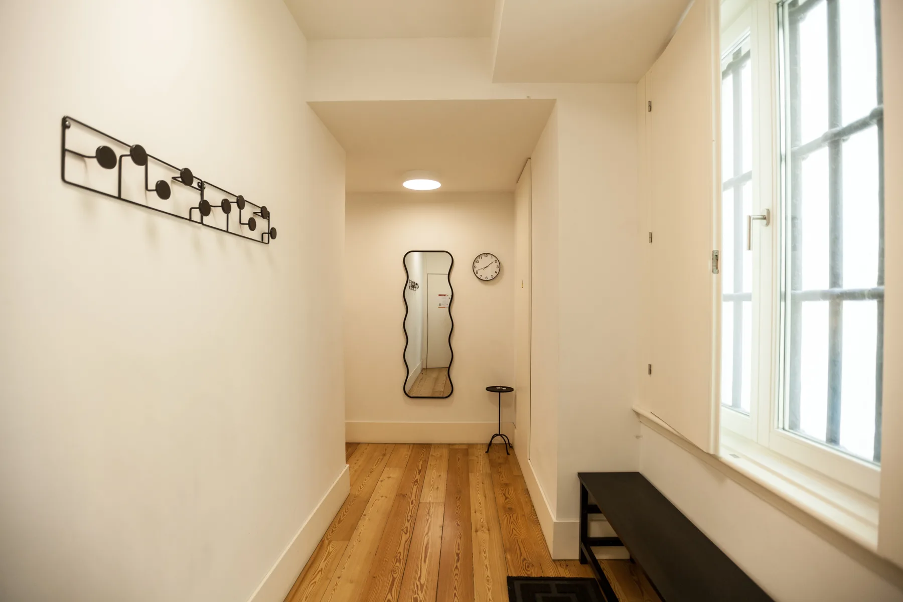 Un pasillo luminoso con suelo de madera, un espejo de pared y ventanas altas.