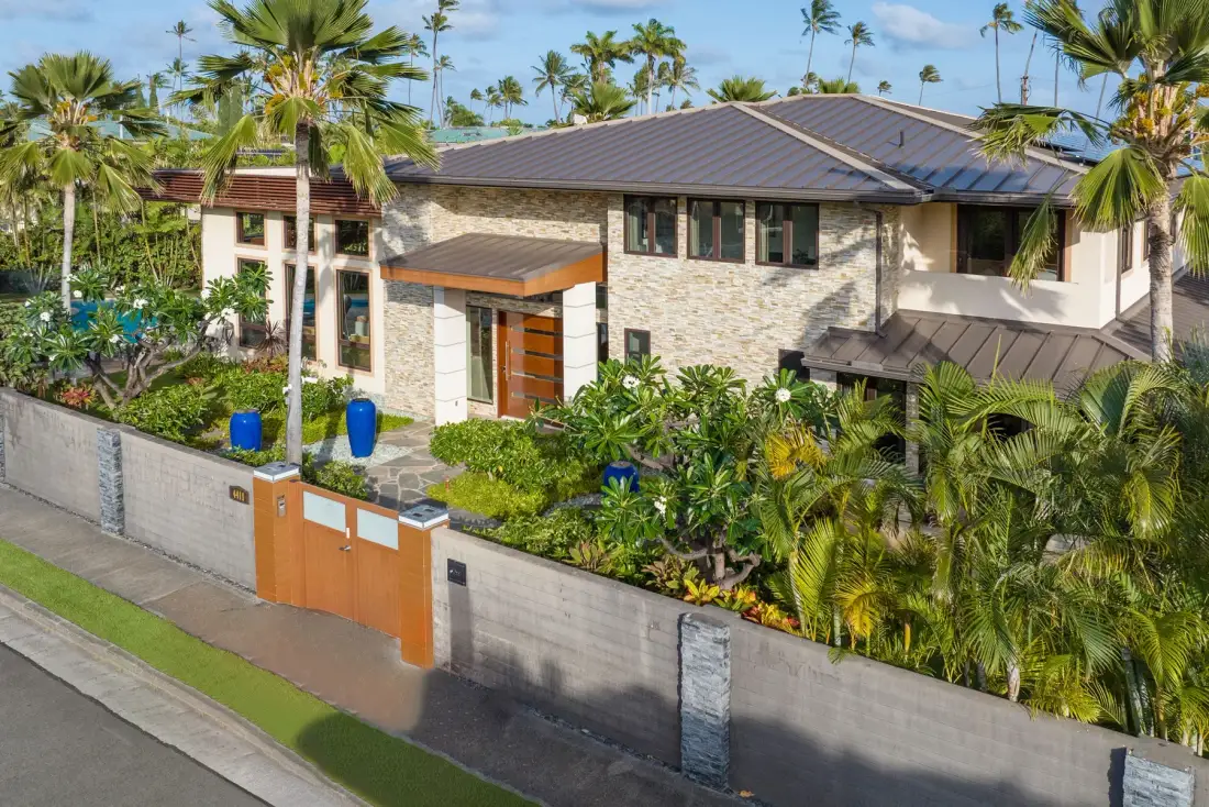 Una casa grande con techo de tejas se encuentra tras un jardín tropical amurallado.