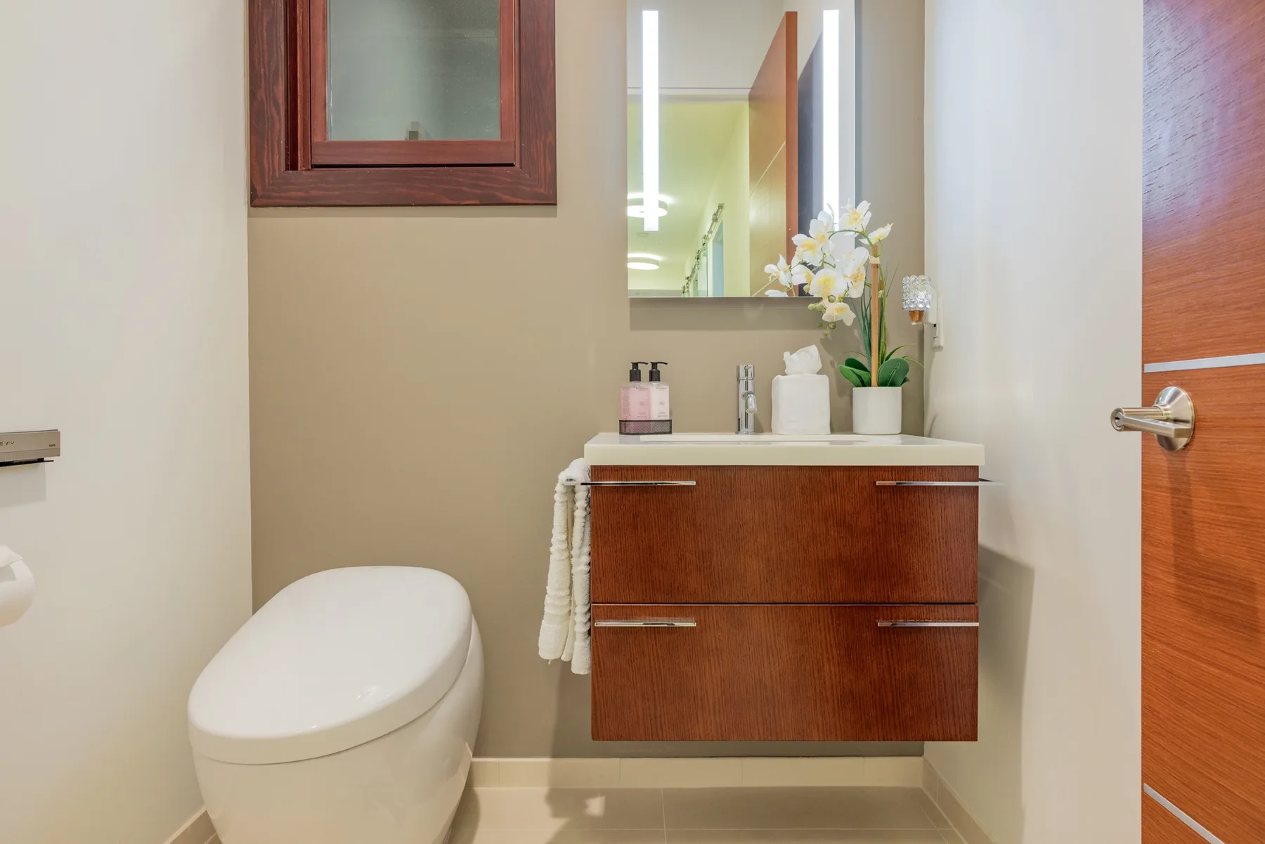 Un baño con un inodoro y un tocador de tono madera bajo un espejo.