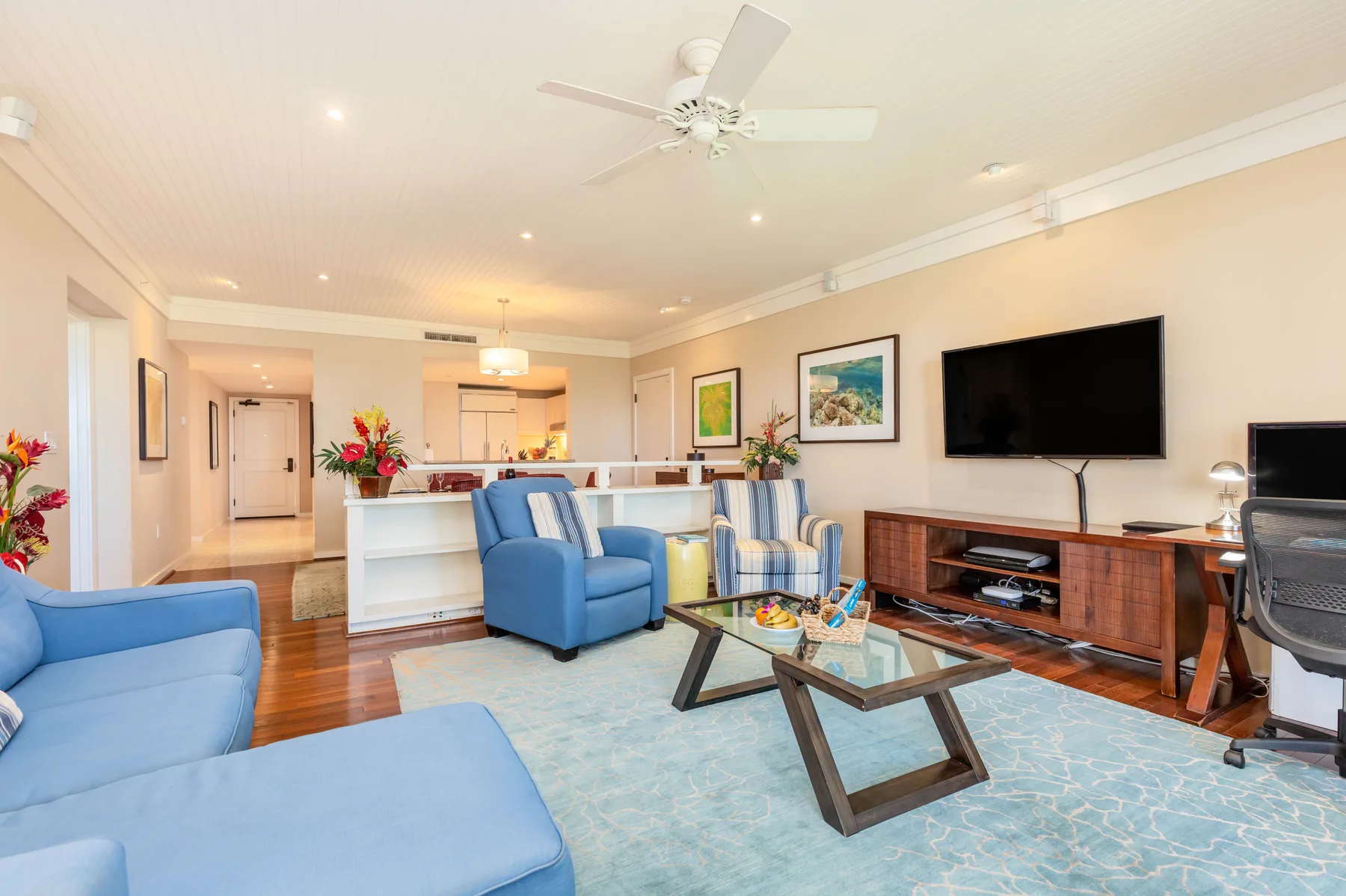 A living room with blue seating, a glass coffee table, and a wall-mounted television.