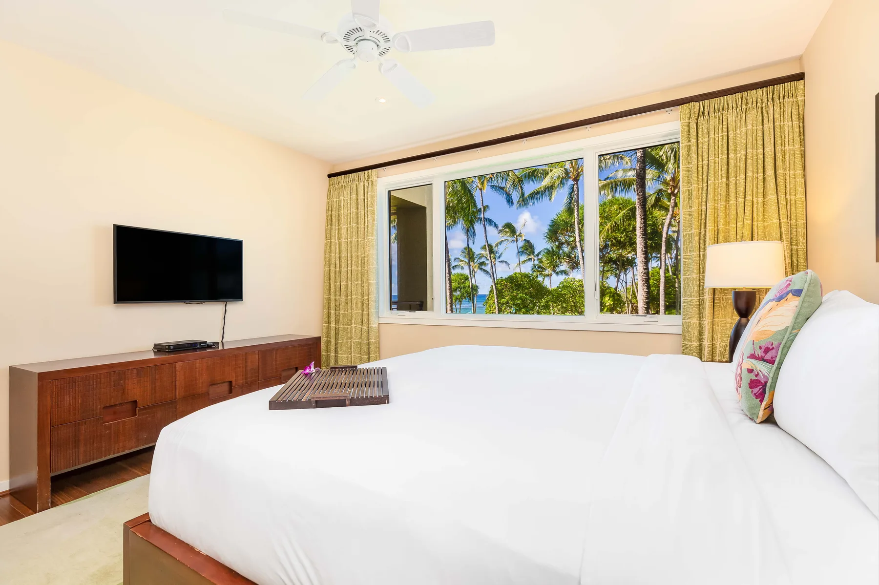A bright bedroom with a large bed facing a window overlooking palm trees.