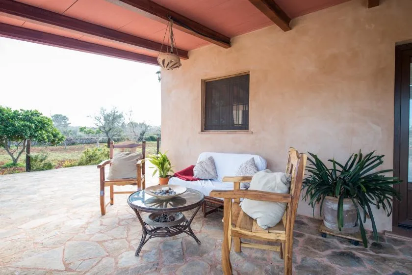 Un patio cubierto con asientos, una mesa baja y plantas junto a un jardín abierto.