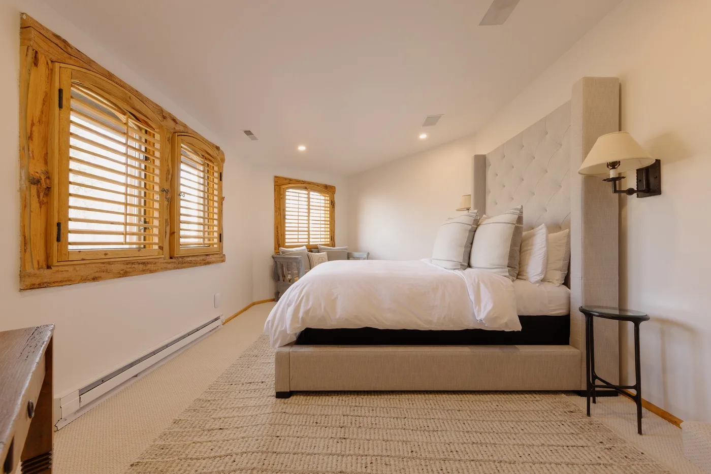 A bright bedroom with a bed, white bedding, and wooden window blinds.