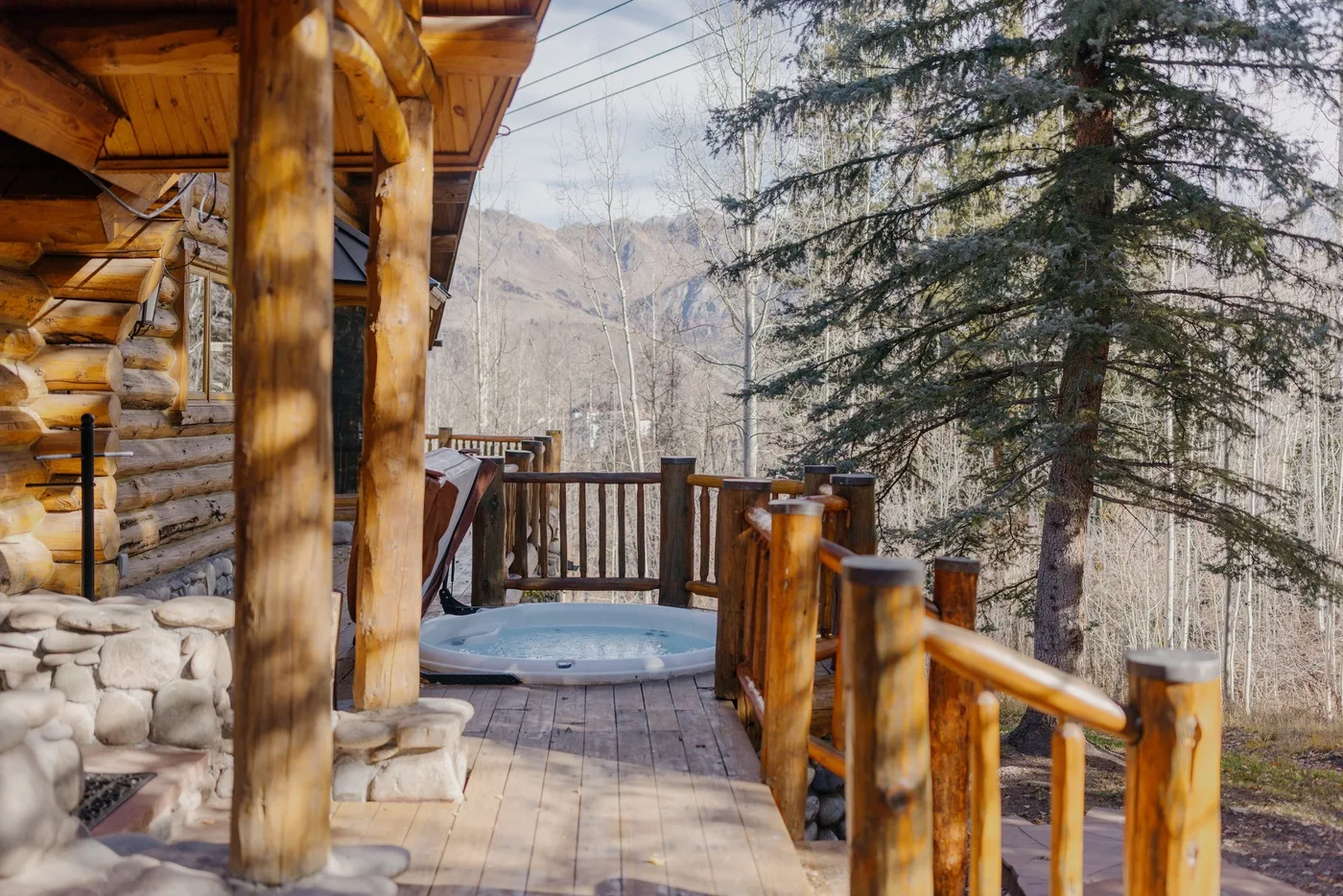 A wooden cabin deck overlooks a hot tub and a wooded mountain landscape.