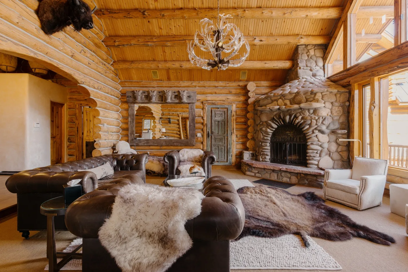 A rustic log-cabin living room has leather seating, a stone fireplace, and large windows.