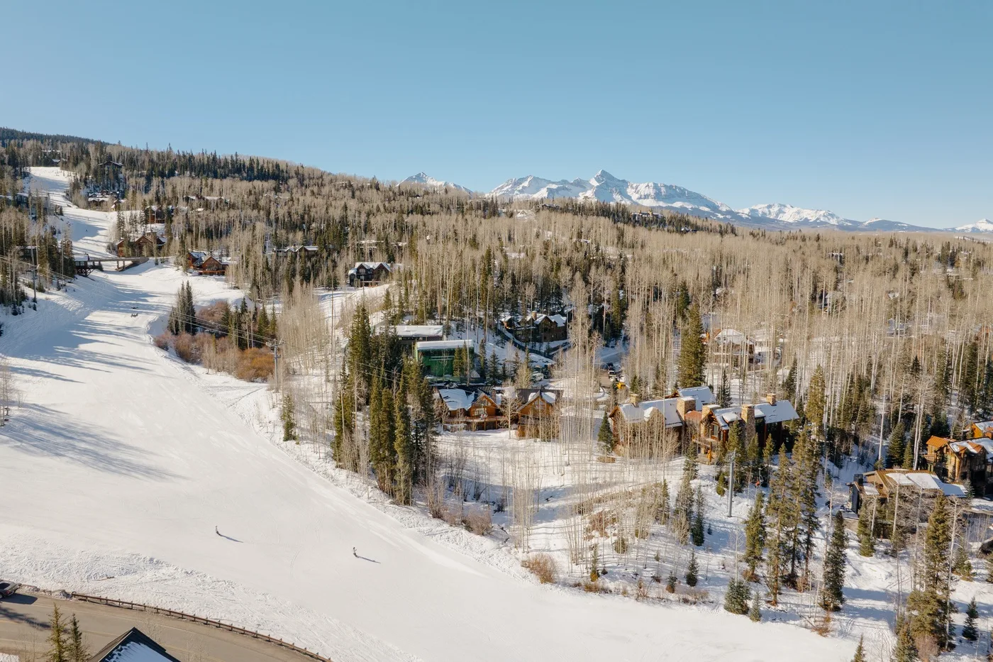 A snowy ski slope runs beside a forested hillside with mountain peaks in the distance.