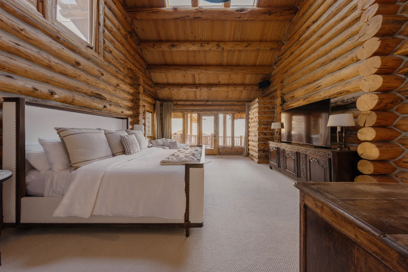 A log cabin bedroom with a bed, a wooden table, and windows overlooking the trees.