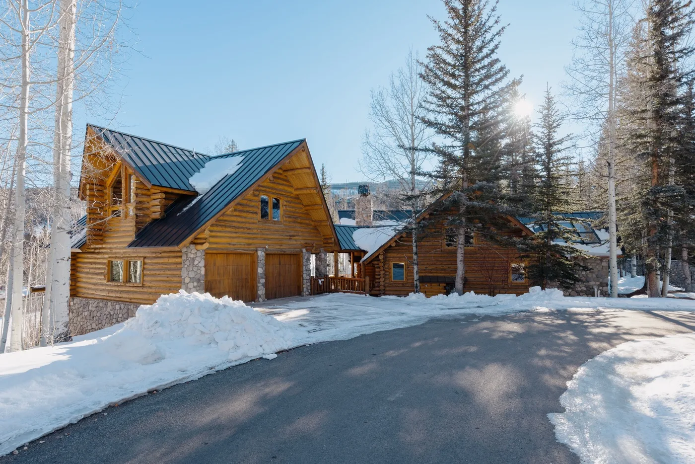 A wooden cabin stands beside a snow-covered driveway among leafless trees.