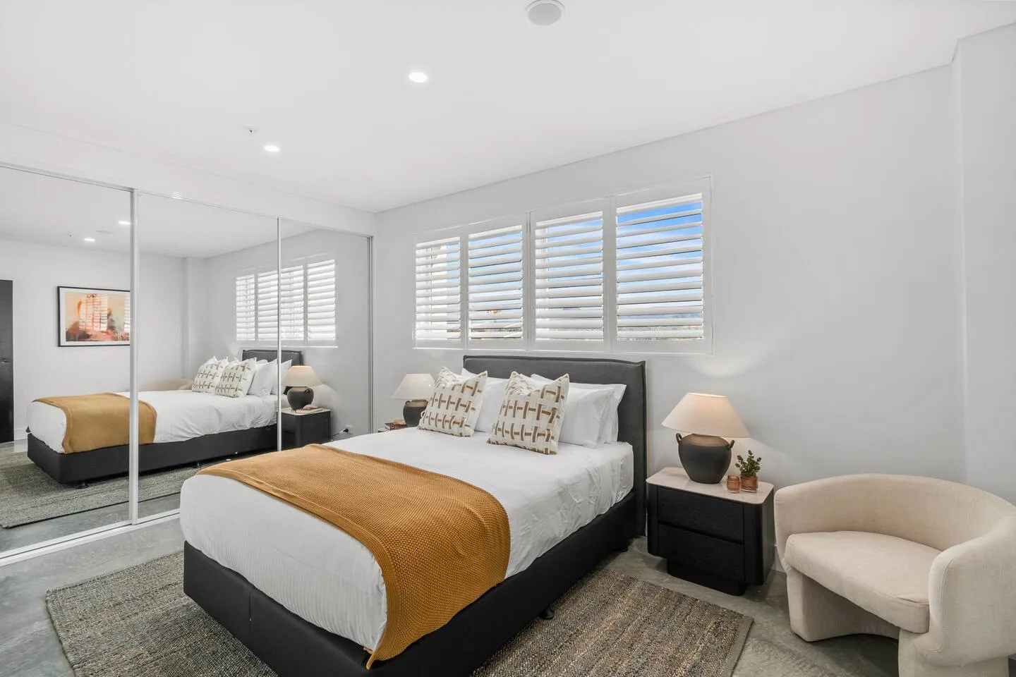 A bedroom with a bed, shuttered windows, a bedside table, and an armchair.