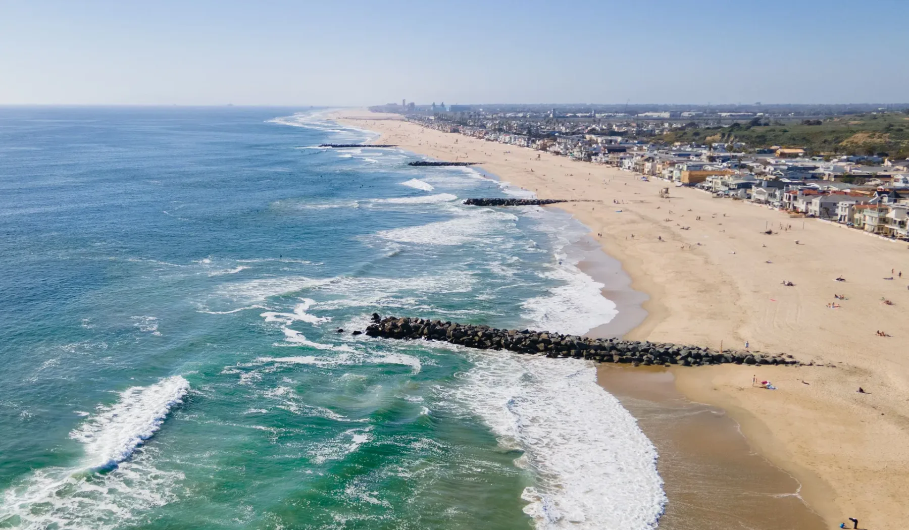 Una vista aérea muestra olas rompiendo junto a una amplia playa de arena y una localidad costera.