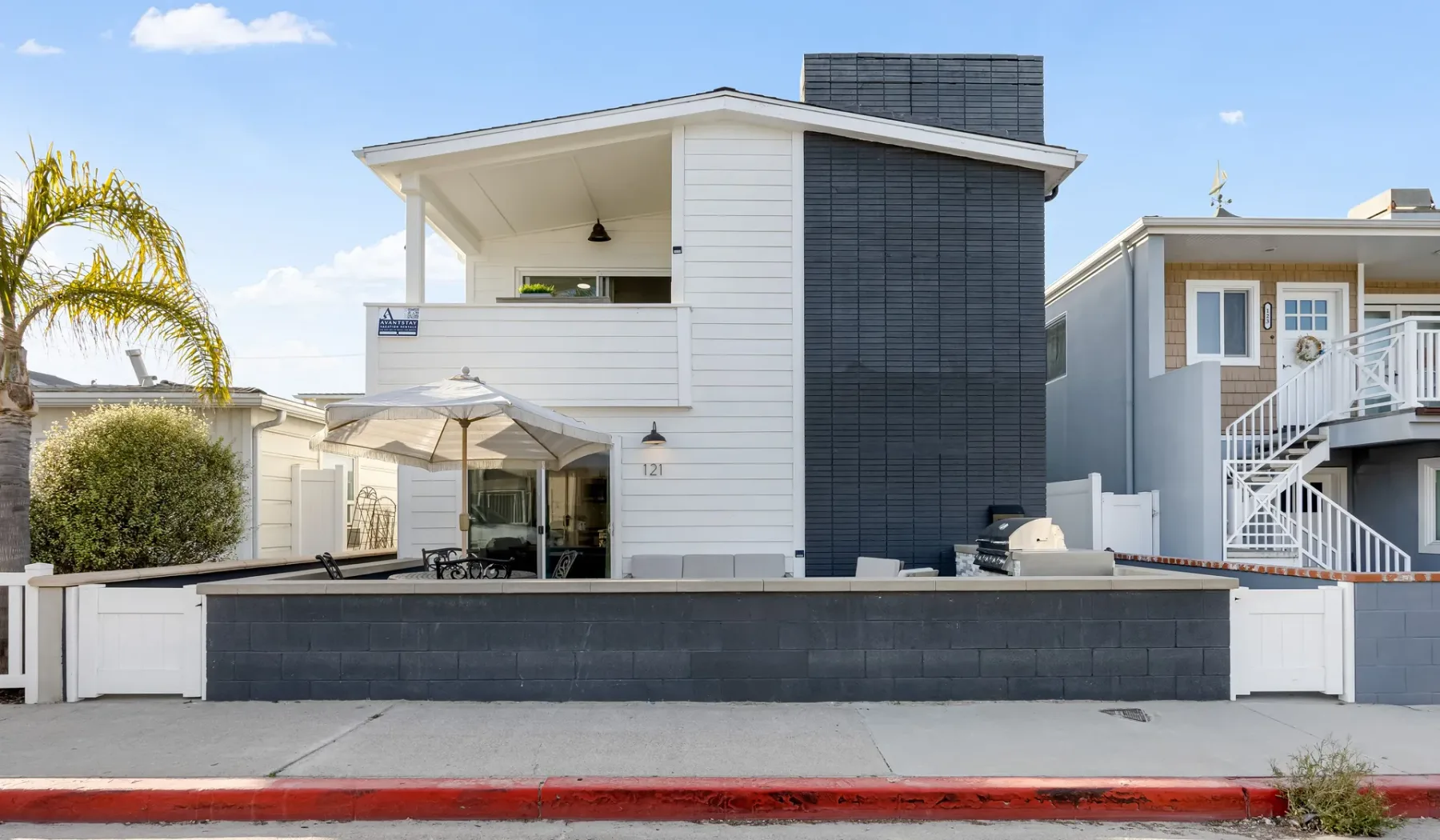 Una casa moderna blanca con una pared exterior oscura y un patio tras un muro bajo.