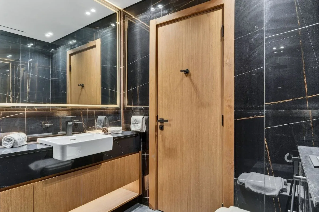 Un baño moderno con una puerta de acabado madera, paredes de azulejos oscuros y un lavabo blanco.