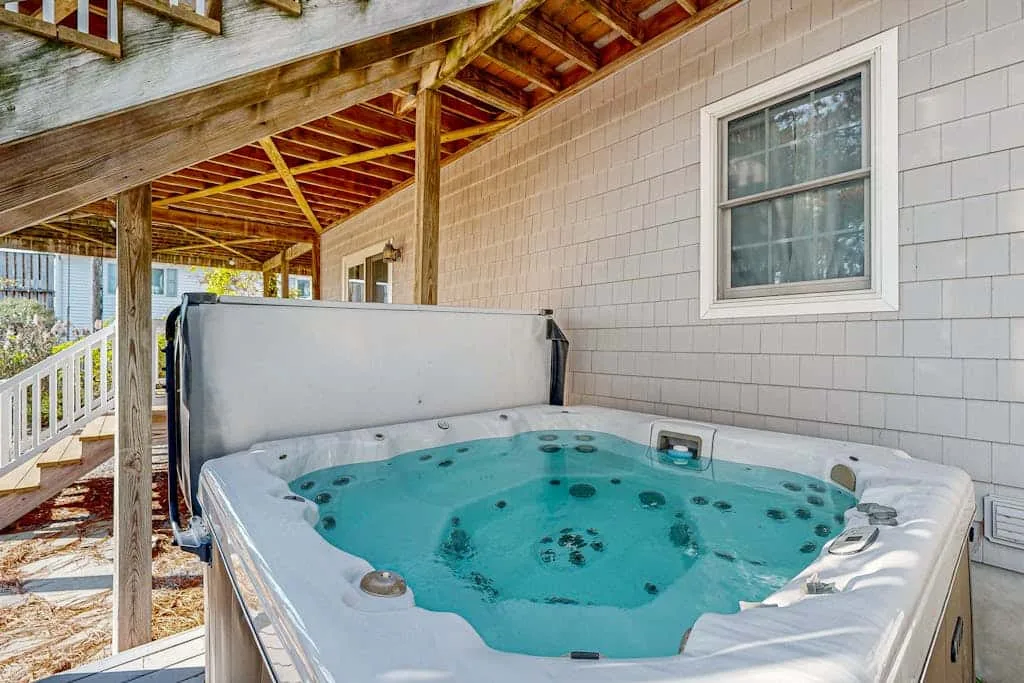 An outdoor hot tub sits beneath a covered wooden deck beside a house.