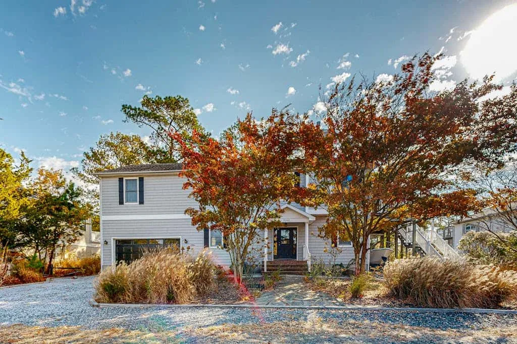 A two-story house stands behind autumn trees beside a gravel driveway.